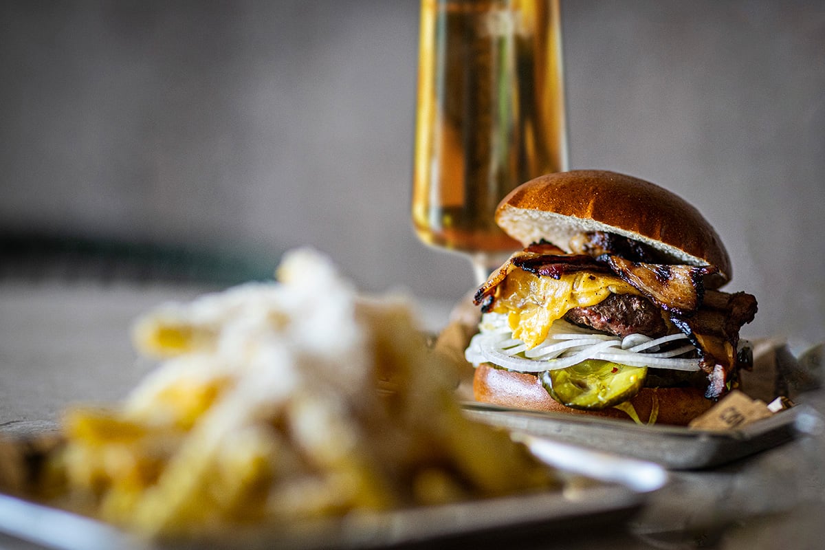 Hamburgare med ost och bacon serverad med pommes frites och ett glas öl i bakgrunden.